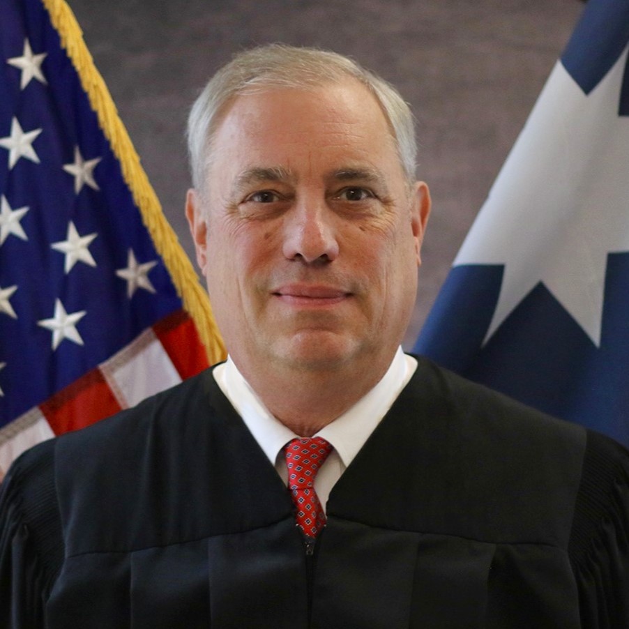 A judge, with silver hair in a white dress shirt, red tie, and black robe, stands in front of a U.S. flag and Minnesota state flag.
