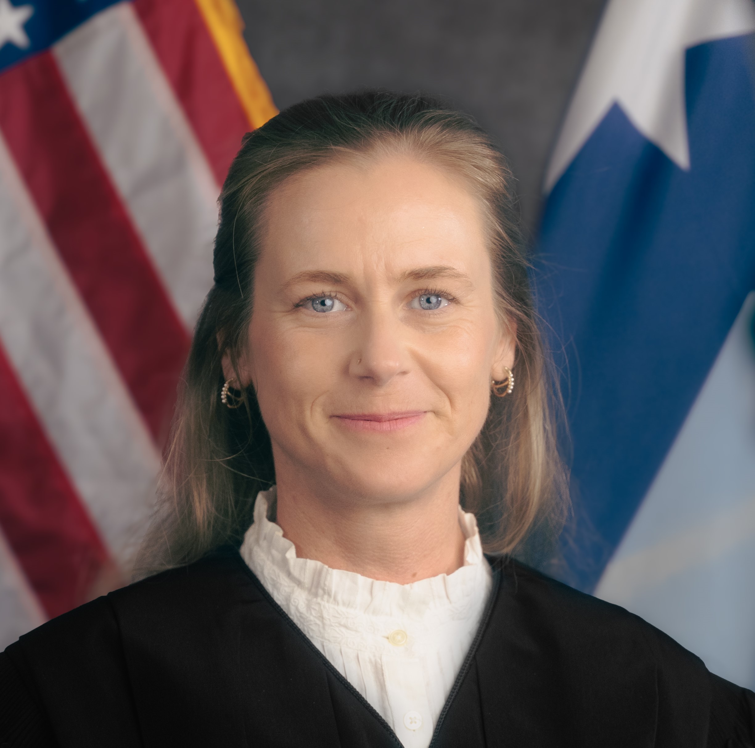 An image of a woman (Judge Jana Leigh) in a judges robe, standing in front of a U.S. flag and a State of Minnesota flag.