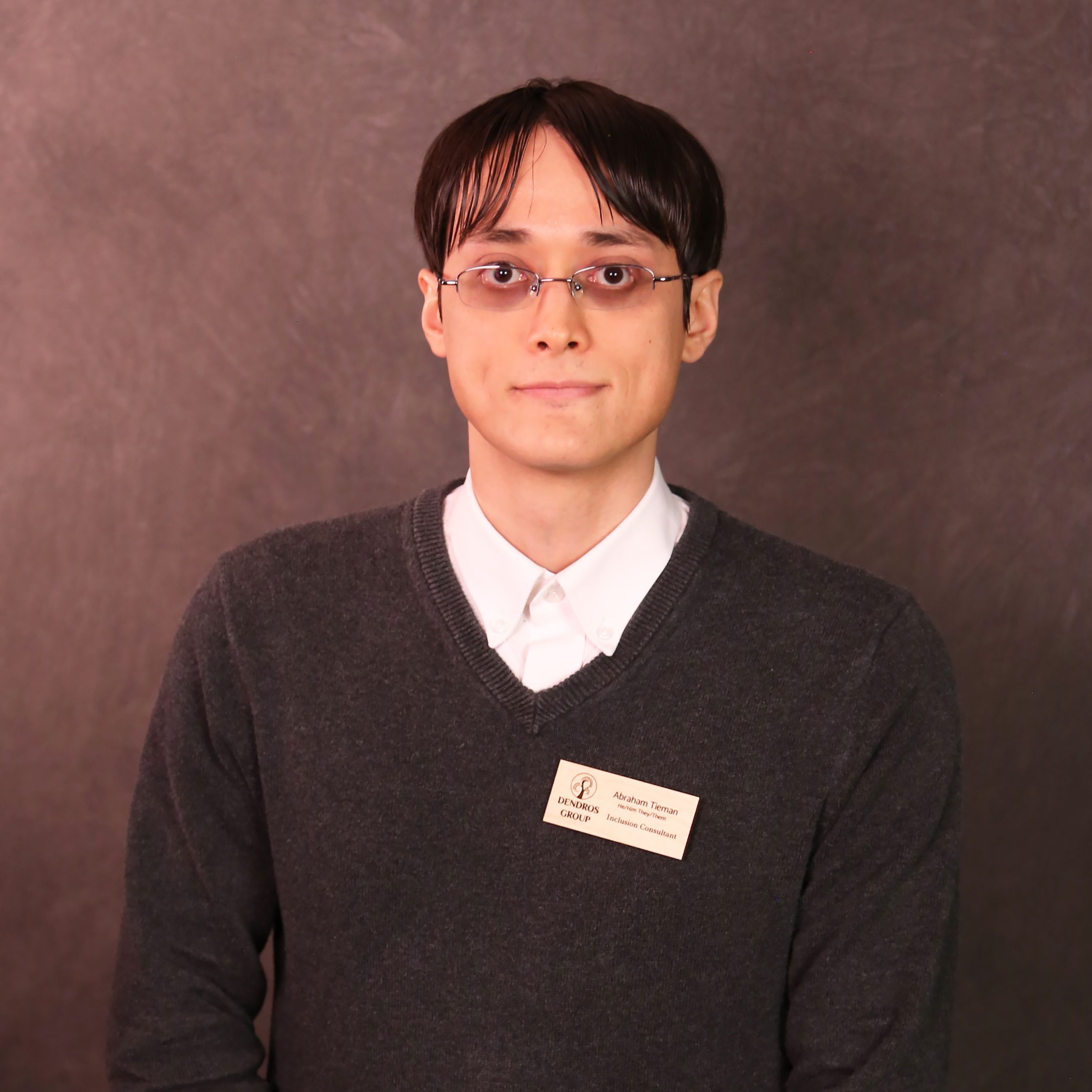 Abraham has tan skin, short dark hair, and wears glasses. They are wearing a white collared shirt underneath a gray sweater.