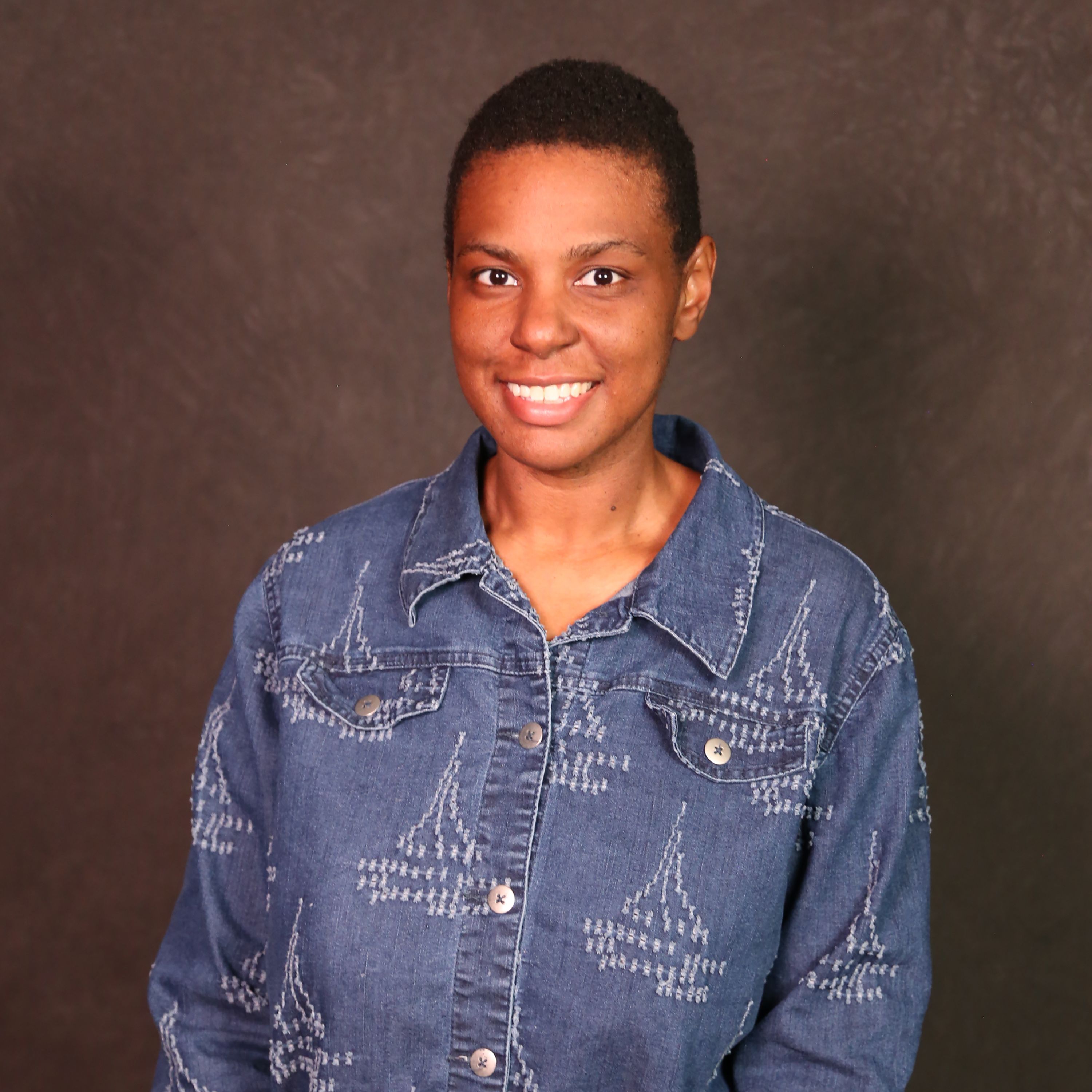Alesha is a Black non-binary person. They have short dark hair and are wearing a denim jacket.