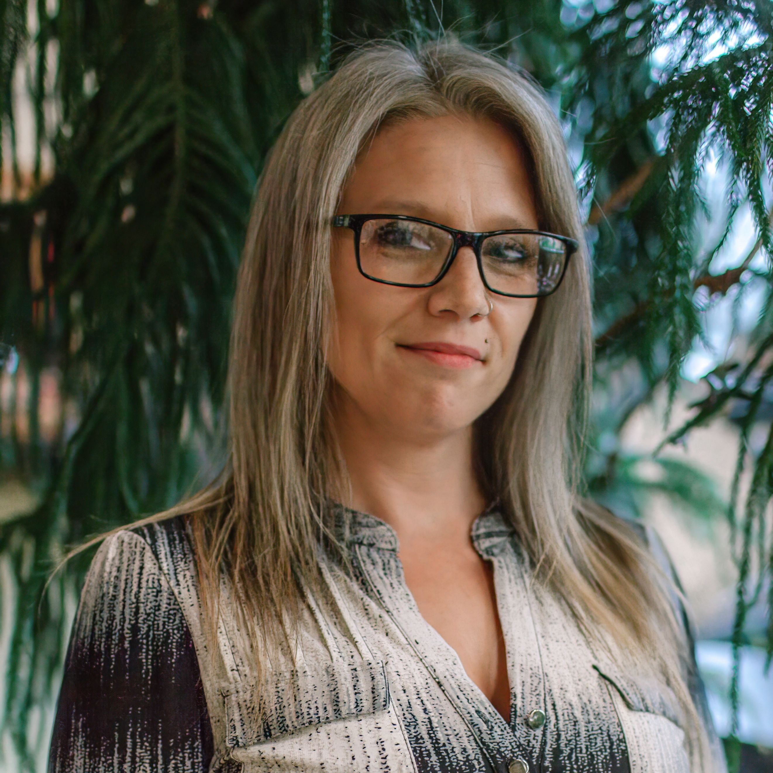 Angela is a woman with dark blonde hair and black frame glasses. She is wearing a black and white button down shirt and is standing outside in front of a pine tree.
