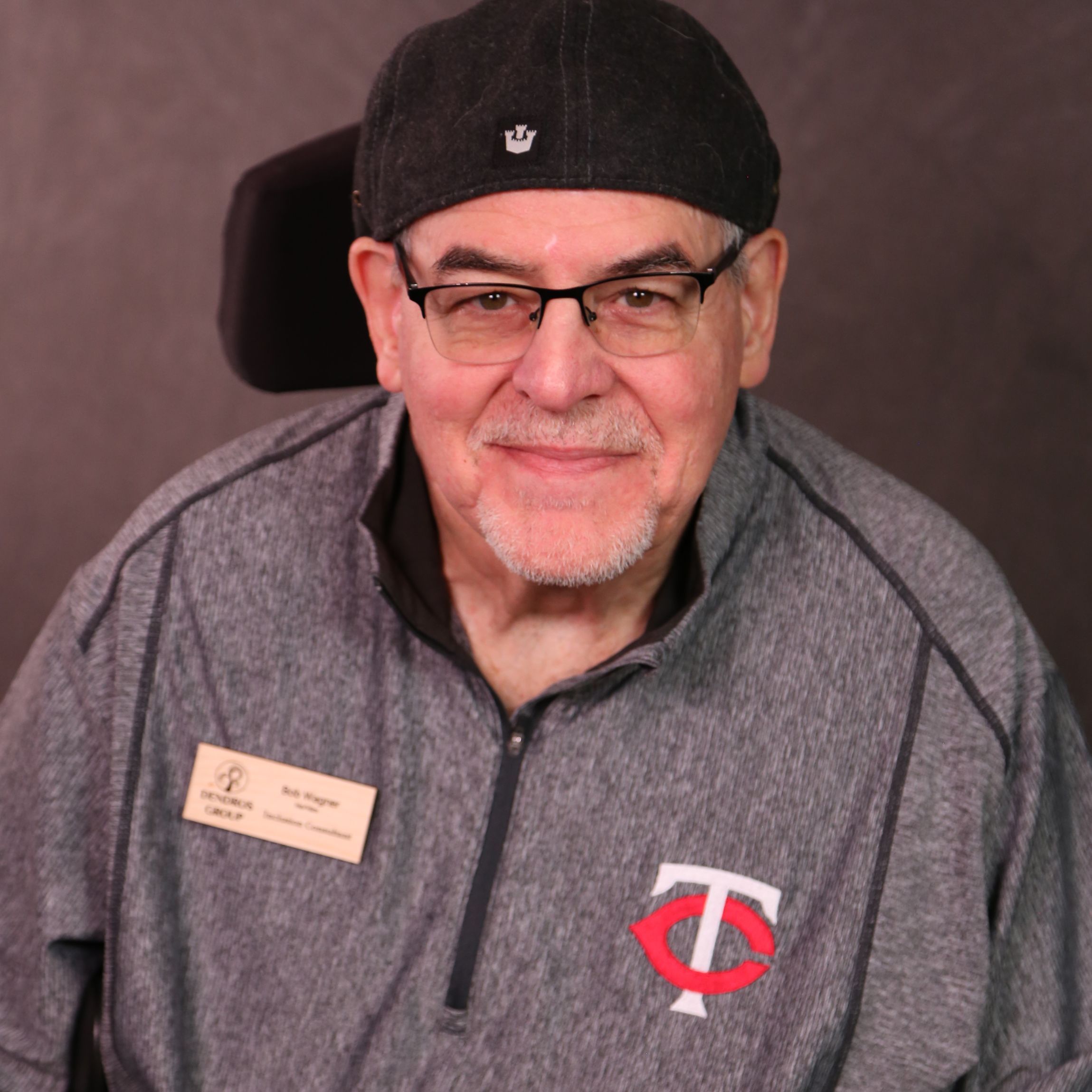Bob is an older man with light skin sitting in a wheelchair. He is wearing a hat, glasses, and a Minnesota Twins shirt.
