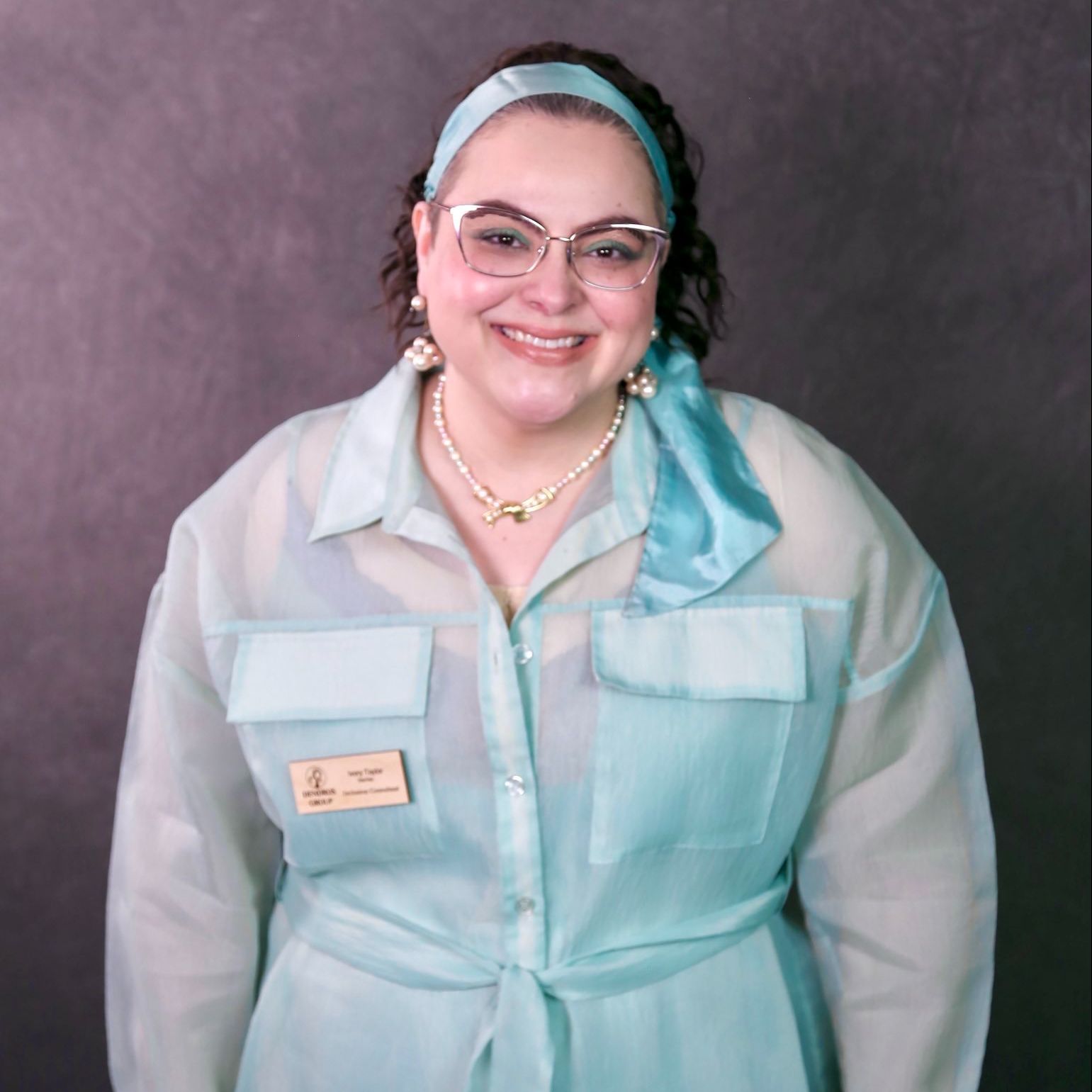 Ivory is a bi-cultural woman with fair skin. She has long curly dark hair pulled back with a headband. She is wearing glasses and a teal dress and fun pearl jewelry.