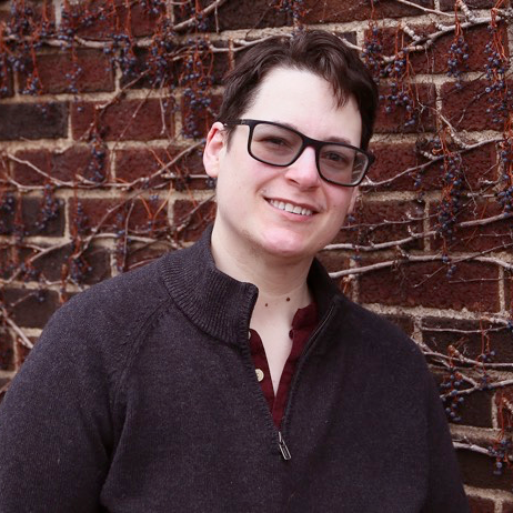  James is a white man with short dark hair and glasses. He is standing in front of a brick wall and wearing a gray sweater.