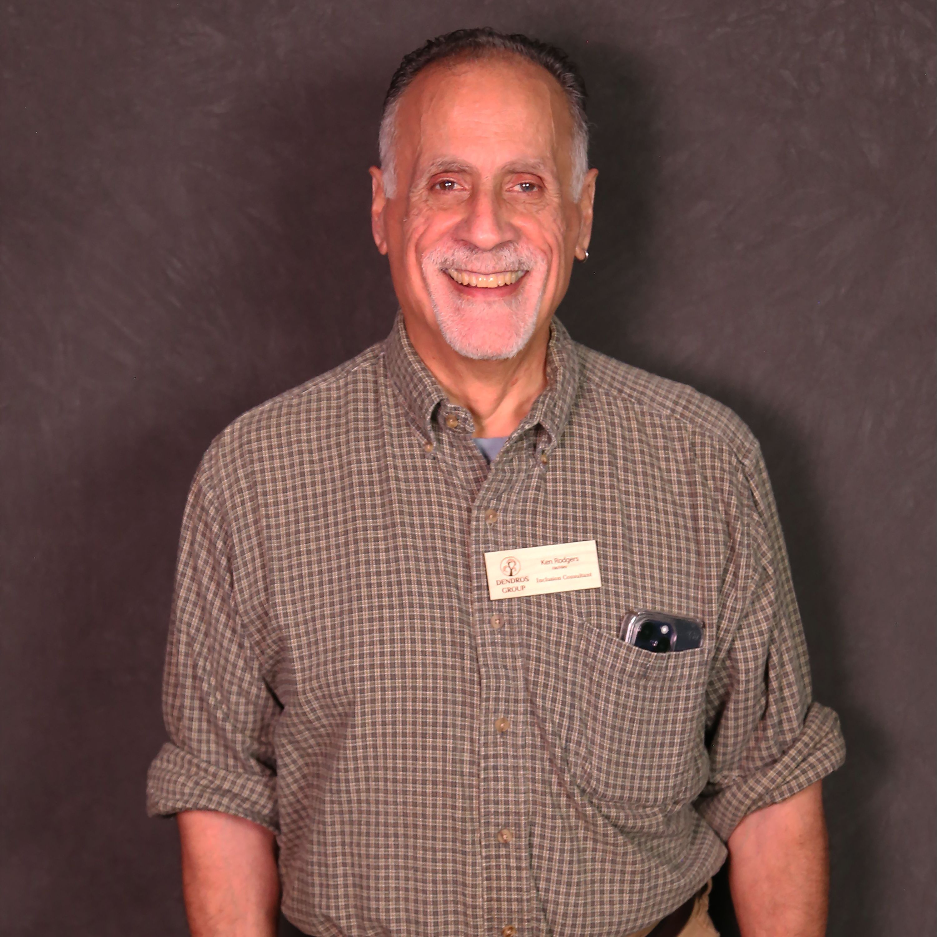 Ken is a man with tan skin, white hair, and a short white beard. He is wearing a brown plaid collared shirt.