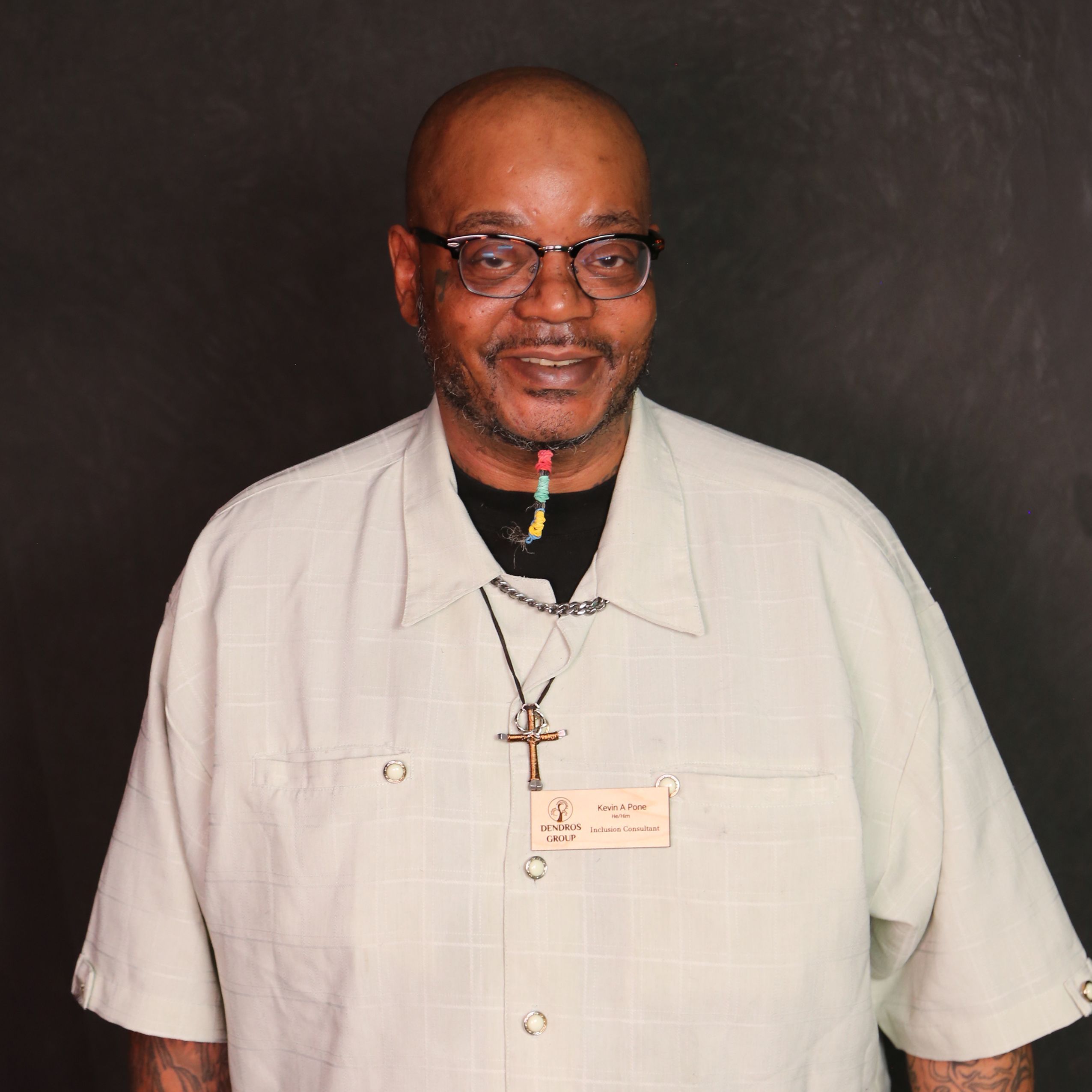 Kevin is a middle aged man with dark skin tone. He is bald and has a goatee that is wrapped in multiple colors. He wears glasses and a tan button down shirt.