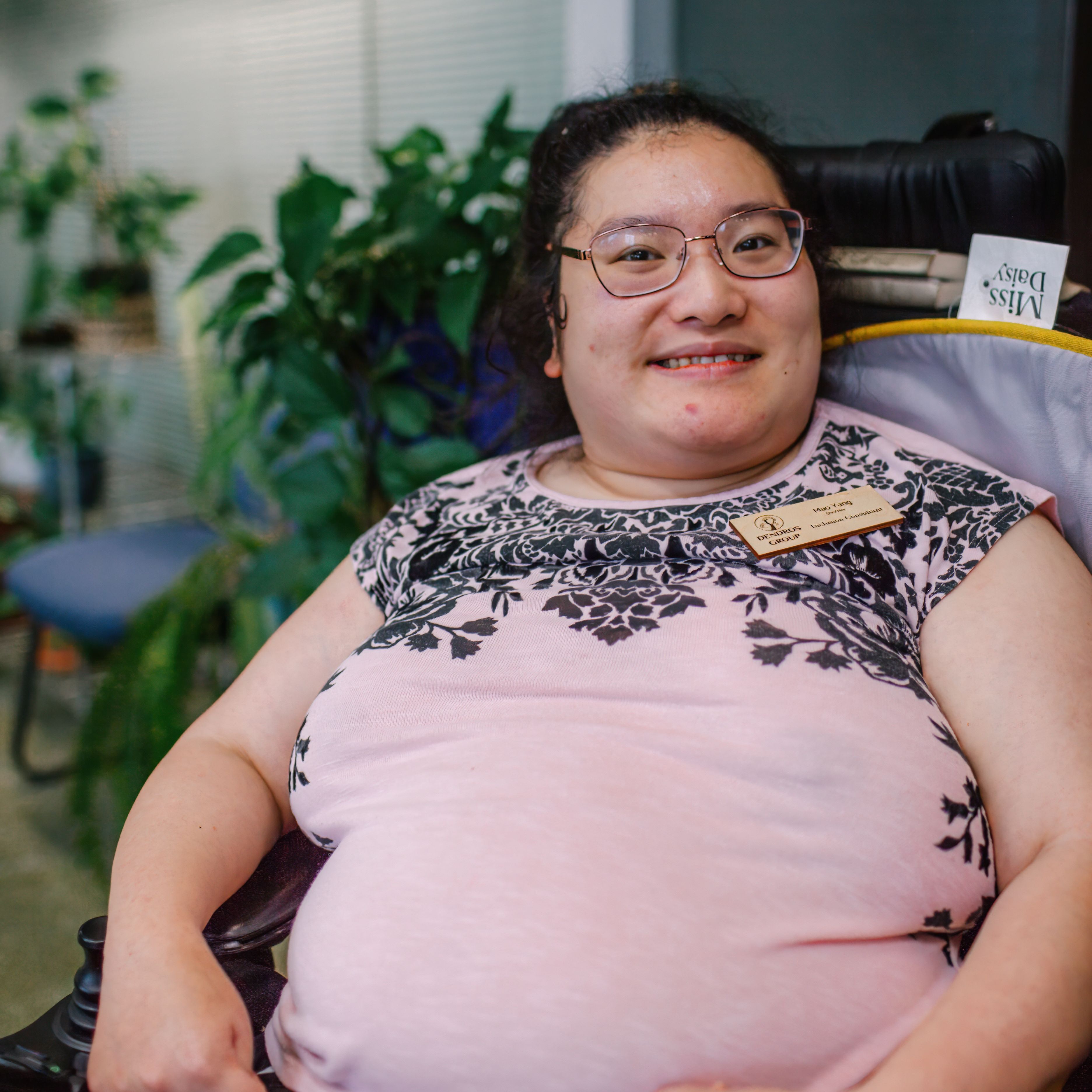 Mao is a young Asian Hmong woman. She is sitting in her powered wheelchair and smiling. She wears a light pink t shirt and has her dark hair pulled back. She is wearing wire rimmed glasses.