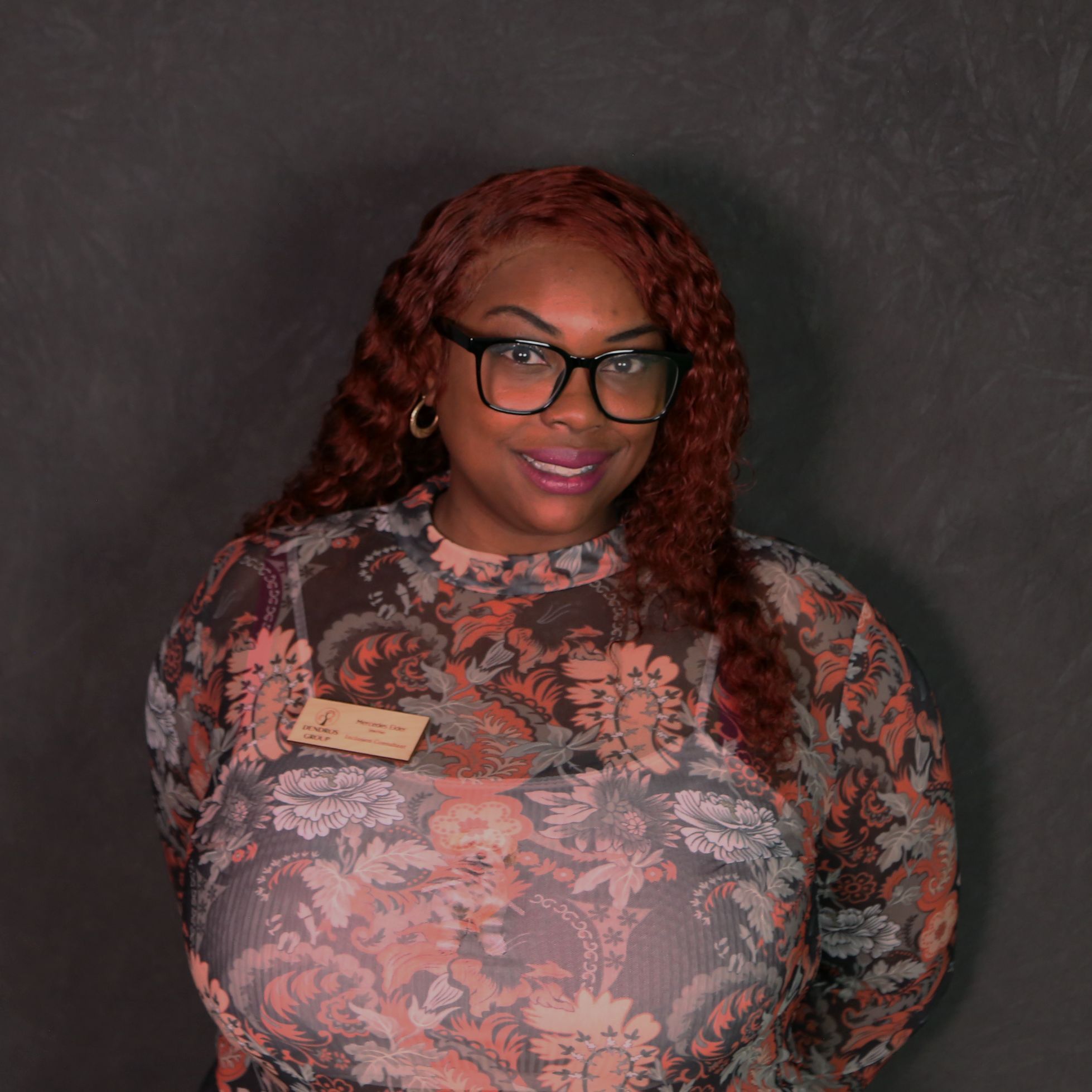 Mercedes is a Black woman with long, wavy, reddish-brown hair. She is wearing glasses and a long-sleeved paisley shirt. 