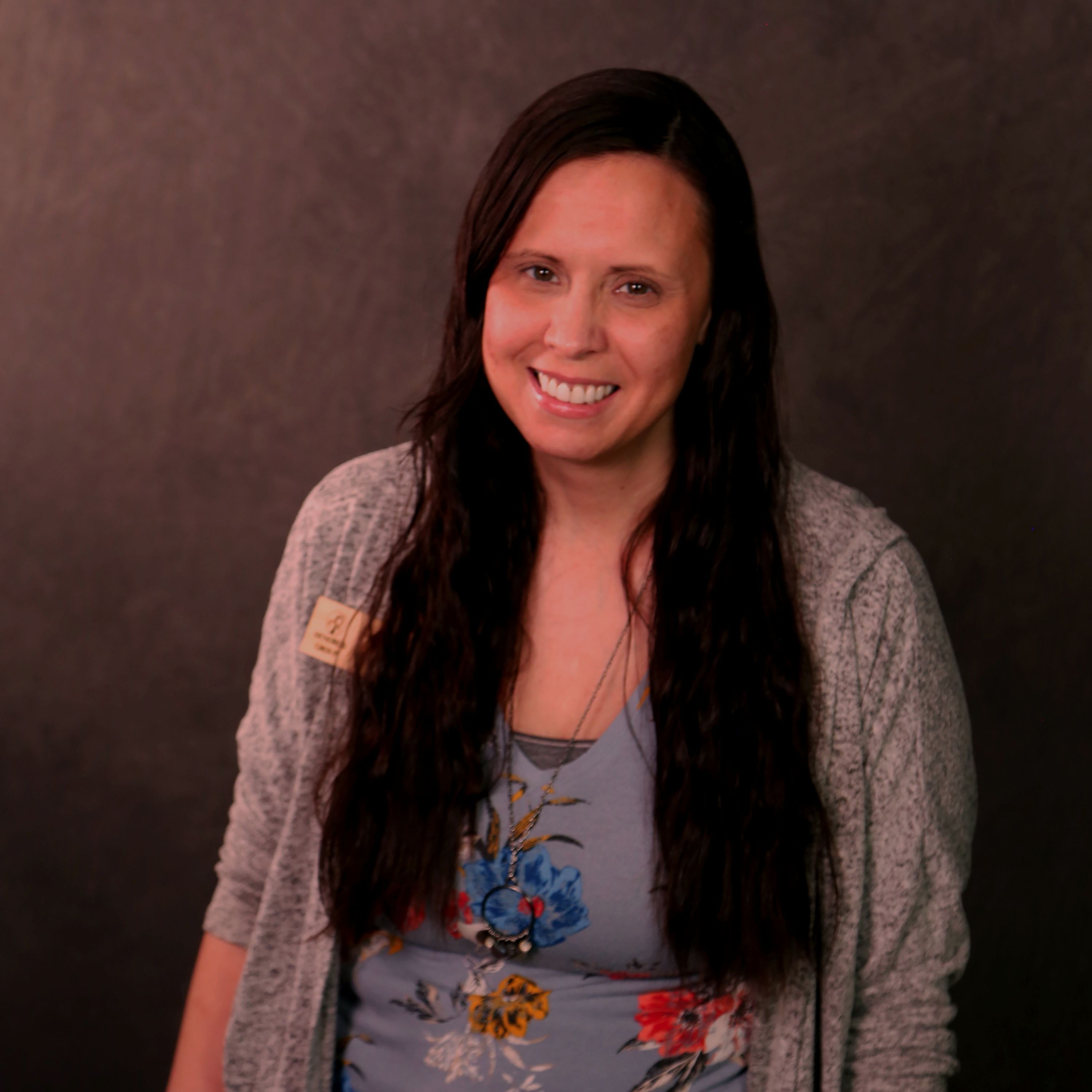Nikki is an Ojibwe woman with long, dark hair. She is wearing a blue floral shirt and a grey cardigan. 