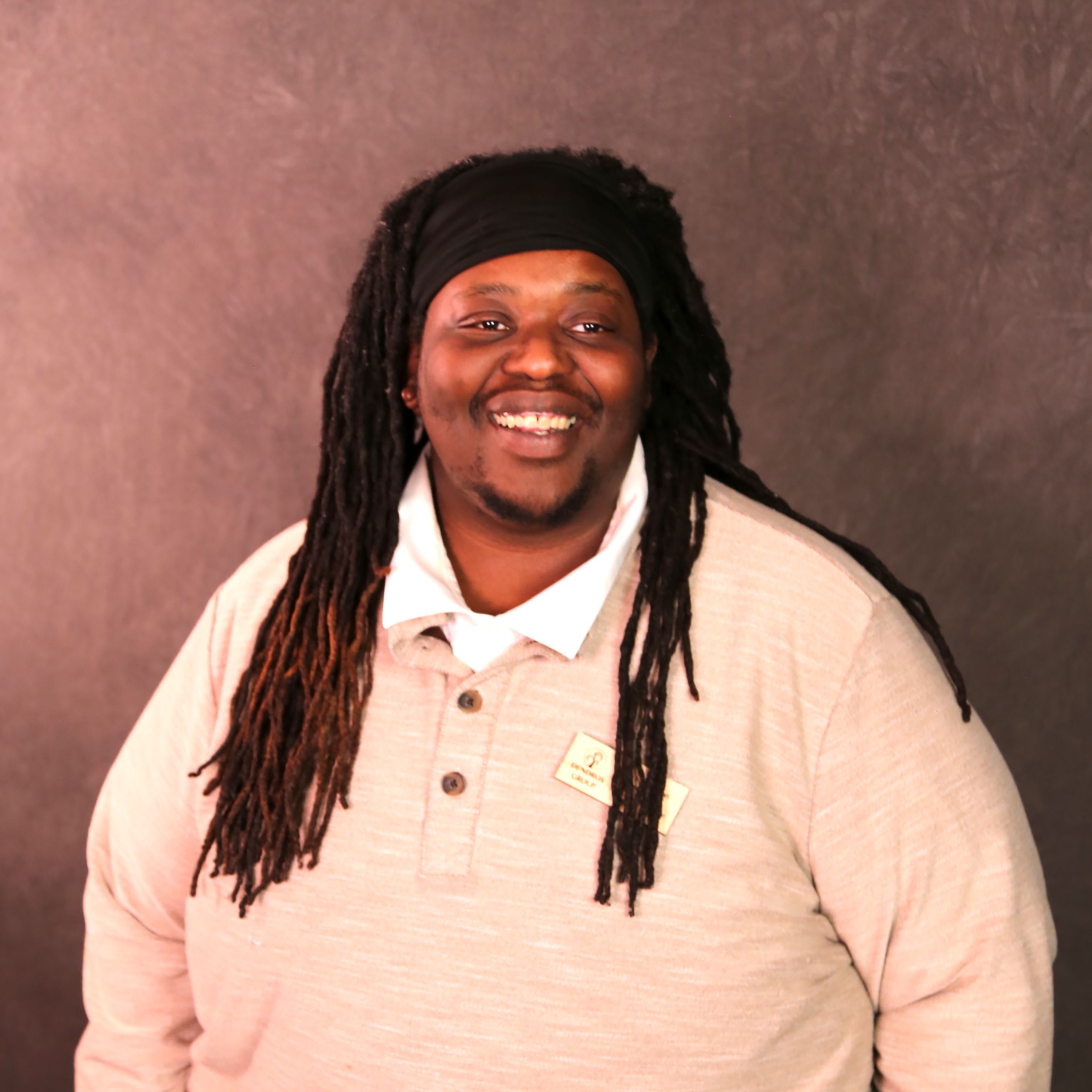 Rich is a Black man with long locs. Rich is wearing a tan shirt with a white collar.