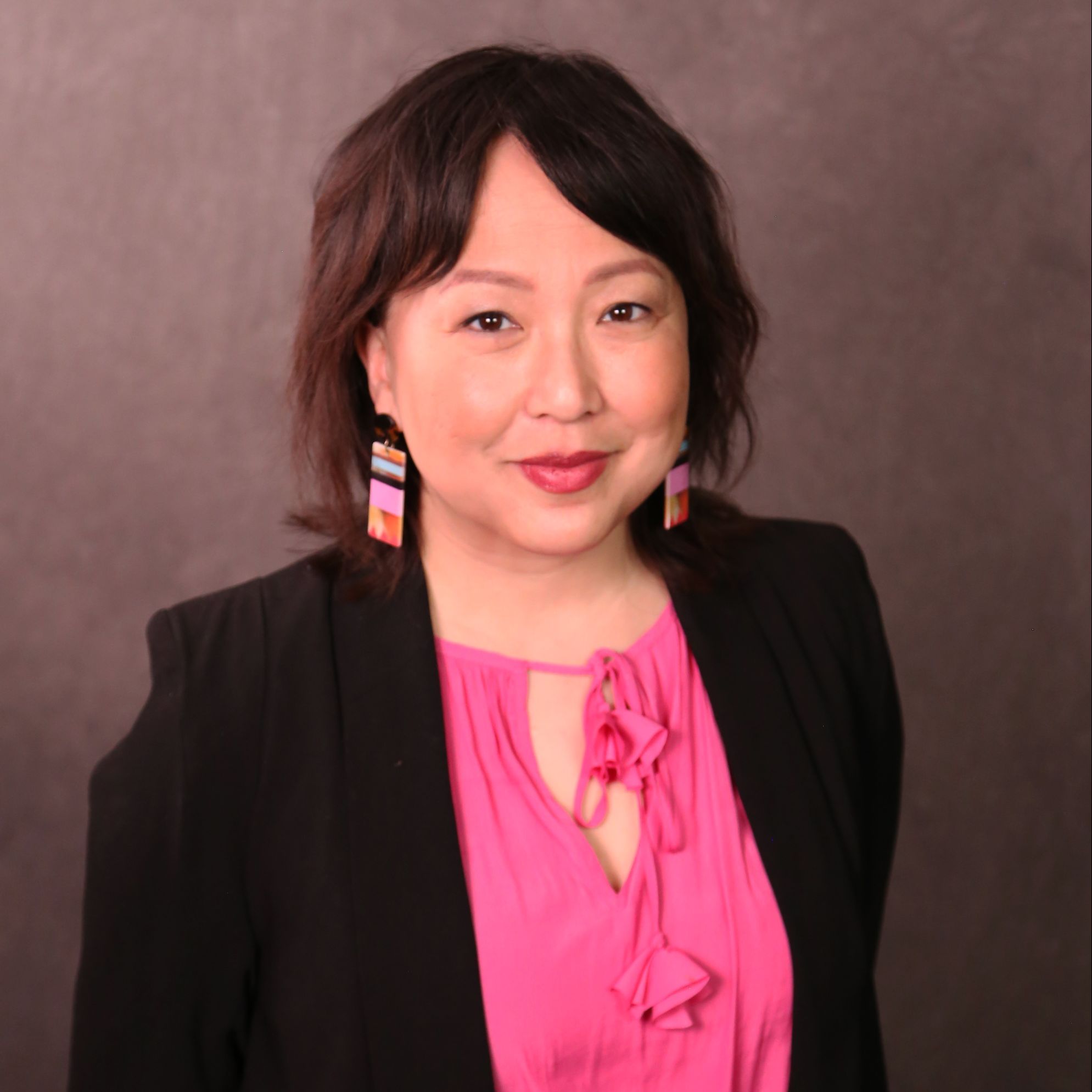 Sandy is a Hmong American woman with wavy brown hair and bangs. She is wearing a black suit with a bright pink shirt and fun earrings.