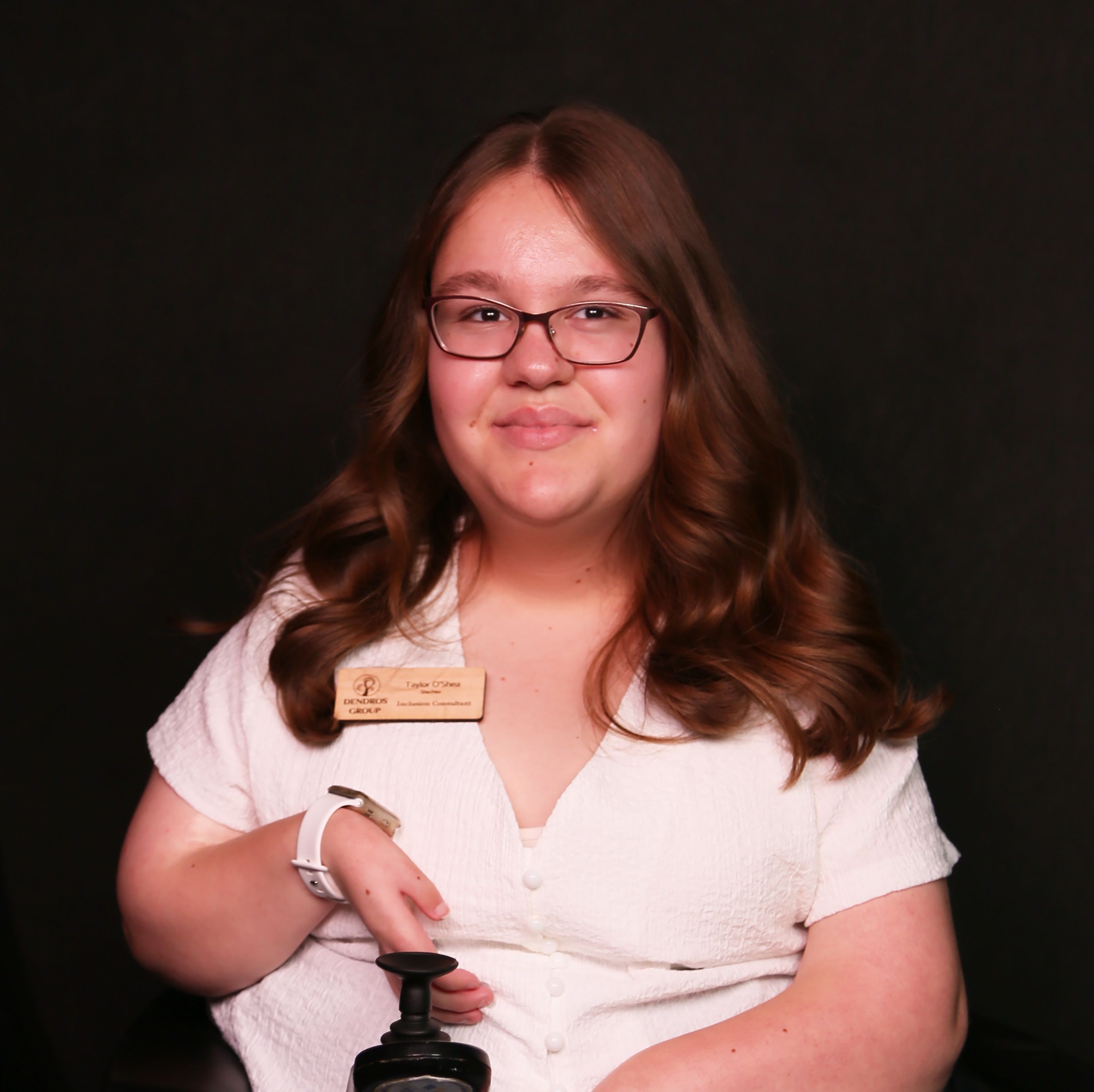 Taylor is a young Ojibwe woman. She has long, medium brown wavy hair and glasses. She is wearing a white shirt and is in her power wheelchair. 