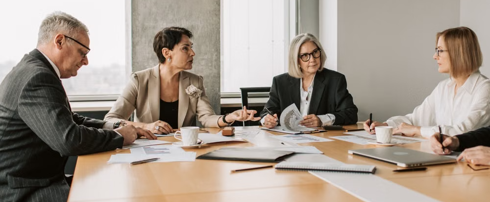 A group of men and women representing a board of directors sit around a table having a meeting and reviewing documentation