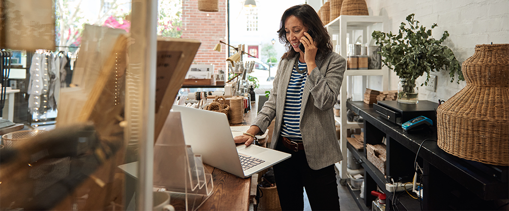 Female small business owner on the phone while looking at her laptop on the counter of her small business. 