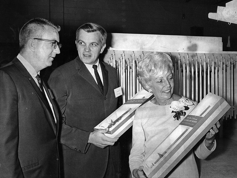 Photo of Mrs. Hubert Humphrey touring a sheltered workshop and holding a packaged set of long candles, with two men dressed in suits standing beside her