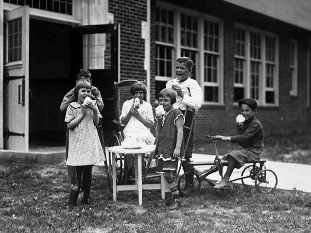 Six children ranging in age from approximately 6 to 10 years eat cake on the lawn of a large brick building.