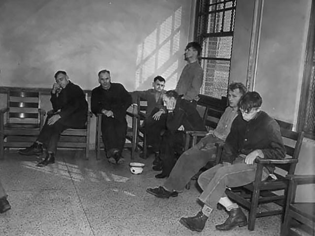 Seven adult males with developmental disabilities sit in benches or stand aimlessly by a window in a large institutional-style room.