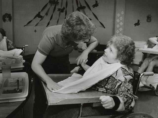 A woman who uses a wheelchair looking up at another woman who is placing a large cloth bib around her client's neck and helping her eat from a lunch tray.