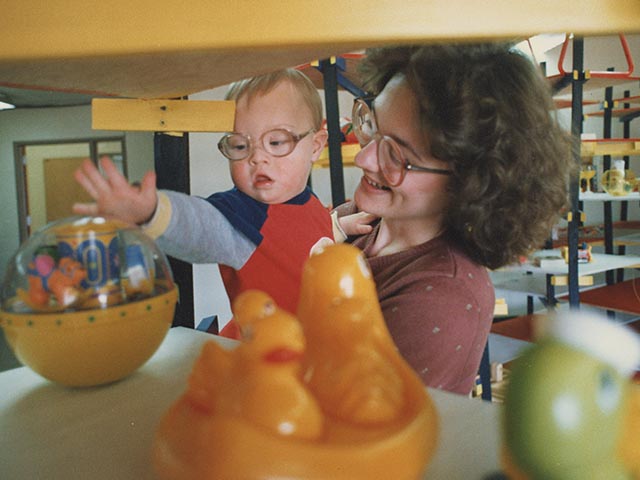 A proud mother holding her baby with Down Syndrome as he reaches for a toy on a shelf.