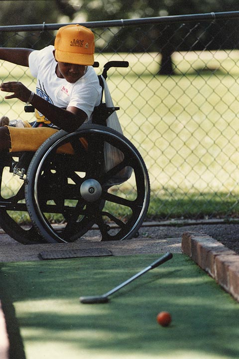 A pre-teen boy reaching for his golf club from his wheelchair.