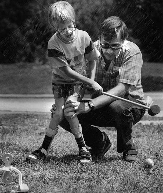 A dad in sunglasses crouching down to support his son's knees as the boy happily swings a croquet mallet.